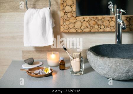 Accessoires de salle de bains naturels : brosse à dents en bambou, démaquillant dans un récipient en verre, bougie de cire de coco, sel de bain biologique, pierre ponce volcanique et serviette en coton près de l'évier en pierre noire. Banque D'Images