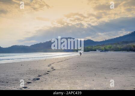 Amérique Centrale, Nicaragua, San Juan Del Sur. Une femme surfant laissant des pieds sur une plage de sable blanc vide au coucher du soleil Banque D'Images