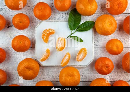 Segments et feuilles de mandarine sur planche à découper blanche entourés de tangerines oranges entières sur une vieille table en bois fissuré. Les fruits et les feuilles sont naturels Banque D'Images