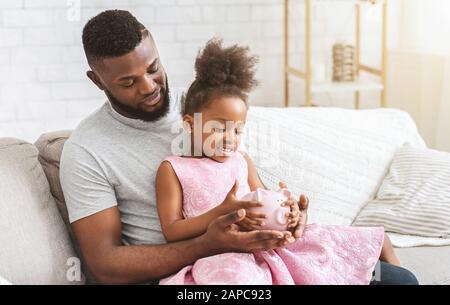 Jeune père afro montrant la petite banque de porc fille Banque D'Images