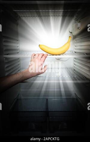 Une main d'homme atteint pour une seule banane illuminant dans le noir dans un réfrigérateur ouvert et vide. Perte de poids concept de régime. Banque D'Images