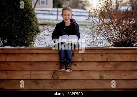 Petit garçon assis sur des planches en bois et lisant la bible dans un jardin couvert de neige Banque D'Images