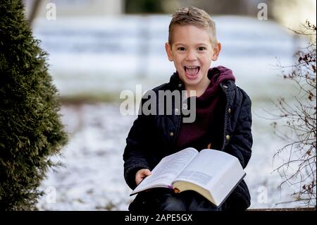 Petit garçon assis sur des planches en bois et lisant la bible dans un jardin couvert de neige Banque D'Images