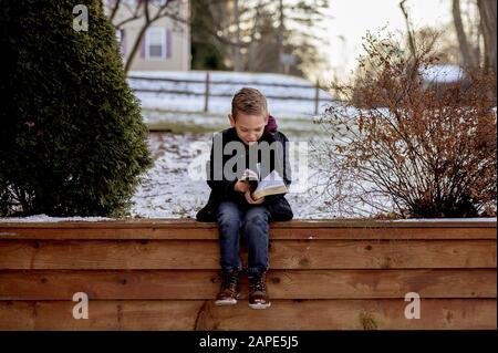 Petit garçon assis sur des planches en bois et lisant la bible dans un jardin couvert de neige Banque D'Images