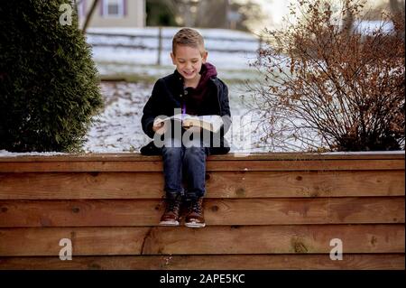 Petit garçon assis sur des planches en bois et lisant la bible dans un jardin couvert de neige Banque D'Images