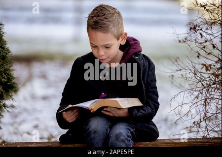 Petit garçon assis sur des planches en bois et lisant la bible dans un jardin couvert de neige Banque D'Images