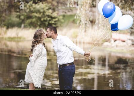 Mari tenant des ballons et embrassant sa femme enceinte dans un jardin avec le lac en arrière-plan Banque D'Images