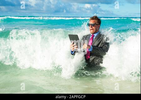 Un homme d'affaires choqué qui tente d'utiliser son ordinateur tablette pour craquer les vagues sur la rive d'une plage tropicale Banque D'Images