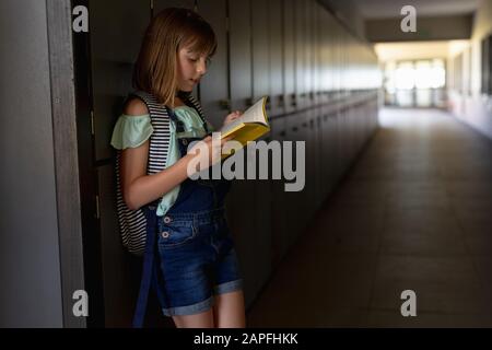 Écolière penchée contre un mur dans un couloir extérieur en lisant un livre à l'école primaire Banque D'Images