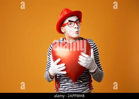 Gros plan m'a surpris dans le chapeau rouge et le maillot de corps tient le ballon de coeur dans ses mains sur fond orange vide Banque D'Images