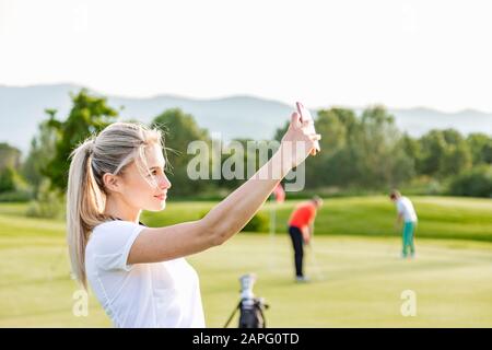 Femme prenant selfie sur le terrain de golf, amis jouant au golf en arrière-plan Banque D'Images