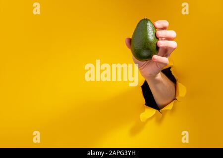 Main donnant un avocat vert à travers un trou déchiré dans fond de papier jaune Banque D'Images