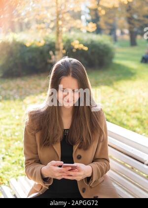 Belle jeune fille assise dans le parc sur un banc, tenant un smartphone et bavardant en ligne sur l'Internet. Femme aux cheveux longs Banque D'Images