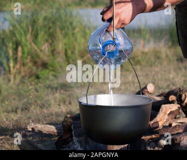 Ajouter de l'eau à partir d'une grande bouteille en plastique dans un chaudron noir sur feu ouvert, repas de camping Banque D'Images