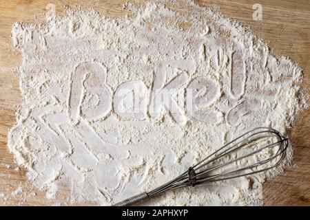 Le mot « Bake! » écrit comme une instruction dans la farine dispersée par l'écriture de doigt sur la planche en bois avec fouet manuel rétro ci-dessous. Banque D'Images