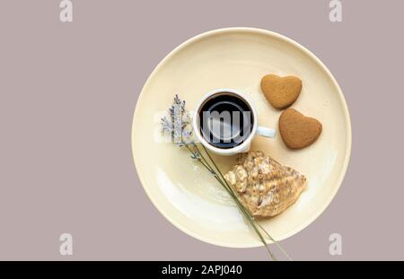 Composition abstraite du petit déjeuner : café, biscuits en forme de coeur, lavande et une coquille sur la plaque jaune pastel isolée sur fond bleu, gros plan, plat l Banque D'Images