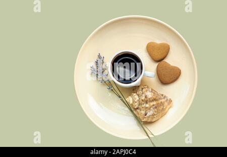 Composition abstraite du petit déjeuner : café, biscuits en forme de coeur, lavande et une coquille sur la plaque jaune pastel isolée sur fond bleu, gros plan, plat l Banque D'Images