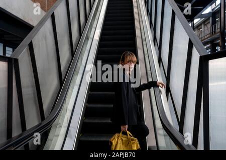 Femme cheveux blonde avec coiffure à demi-égalité debout sur l'escalator tenant le sac à dos jaune prop sa main sur la main courante. Banque D'Images