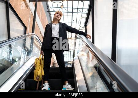 Femme cheveux blonde dans des vêtements semi-formels debout avec de larges jambes sur escalator tenant le sac à dos jaune et reposer son bras sur la main courante, tir à faible angle. Banque D'Images