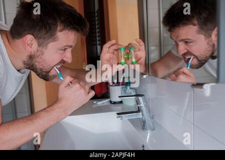 Jeune homme avec dents de nettoyage de brosse à dents et regardant le miroir à la maison salle de bains vérifier l'heure sur l'horloge spéciale. Banque D'Images