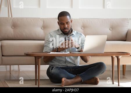 Le gars africain est assis à plusieurs pattes sur la moquette à l'aide d'un smartphone Banque D'Images