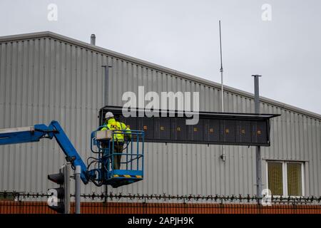 Un employé de maintenance dans des vêtements haute visibilité un chapeau de dur travaillant sur la fixation d'un panneau sur un sélectionneur de cerisier Banque D'Images