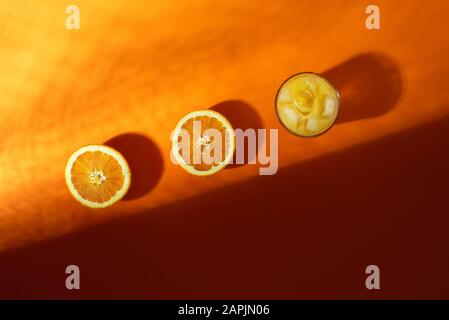 Jus d'orange frais dans un verre avec des glaçons et des fruits d'orange coupés en deux parties sur fond de lave luxuriant, en plein soleil et ombre. Boisson froide d'été. Banque D'Images