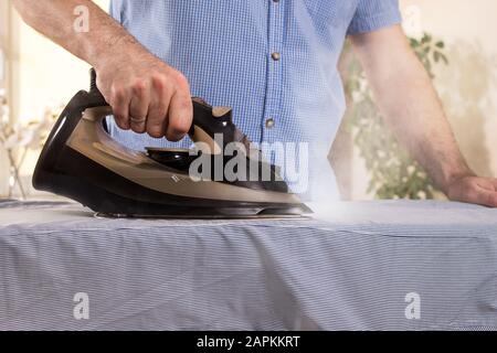 Une main d'homme tient un fer noir et fer à repasser la chemise sur une planche à repasser. Tâches ménagères exercées par des hommes. Banque D'Images