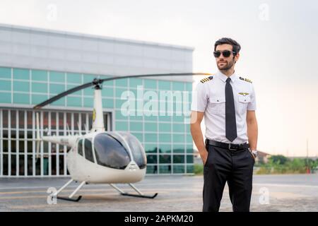 Portrait de beau pilote commercial en uniforme blanc capitaine debout et regardant intelligent près d'un petit hélicoptère privé sur un point d'atterrissage avec privé Banque D'Images