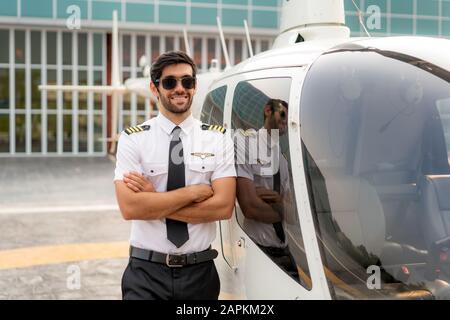 Portrait de beau pilote commercial en uniforme blanc capitaine debout et regardant intelligent près d'un petit hélicoptère privé sur un point d'atterrissage avec privé Banque D'Images