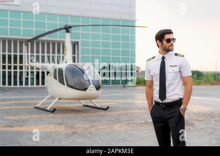 Portrait de beau pilote commercial en uniforme blanc capitaine debout et regardant intelligent près d'un petit hélicoptère privé sur un point d'atterrissage avec privé Banque D'Images