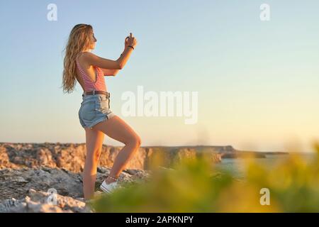 Jeune femme debout sur la falaise en mer, à l'aide d'un smartphone Banque D'Images