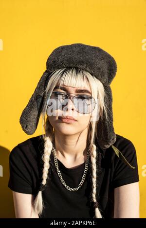 Portrait de la jeune femme avec des cheveux teints portant une casquette et des lunettes de soleil contre le mur jaune Banque D'Images