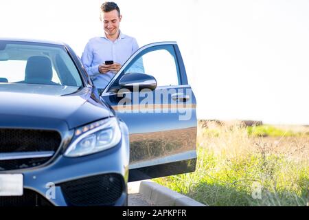 Un jeune homme d'affaires souriant utilisant un smartphone en voiture sur la route nationale Banque D'Images