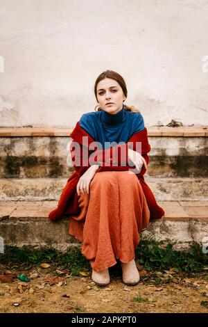 Gros plan portrait d'une femme avec pullover à col roulé bleu et manteau rouge assis sur une marche Banque D'Images