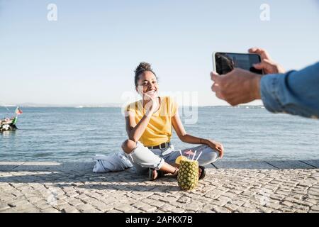 Jeune homme prenant une photo de sa petite amie assis sur la jetée au bord de l'eau, Lisbonne, Portugal Banque D'Images