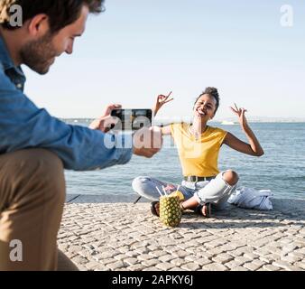 Jeune homme prenant une photo de sa petite amie assis sur la jetée au bord de l'eau, Lisbonne, Portugal Banque D'Images