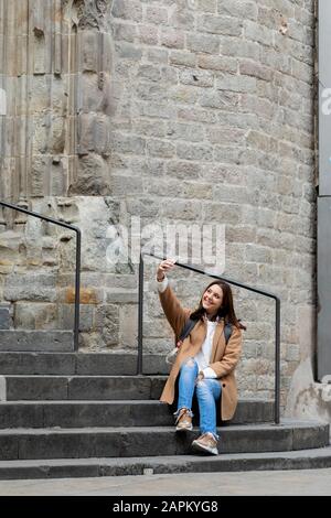 Jeune femme souriante assise dans les escaliers de la ville en prenant un selfie, Barcelone, Espagne Banque D'Images