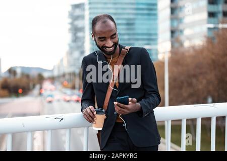 Un jeune homme d'affaires souriant avec du café pour écouter de la musique avec des écouteurs et un smartphone Banque D'Images