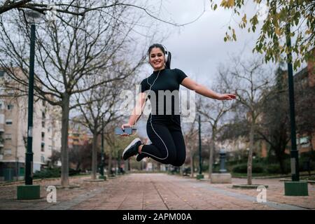 Portrait de la jeune femme dans des vêtements de sport noirs sautant dans l'air tout en écoutant de la musique avec des écouteurs Banque D'Images