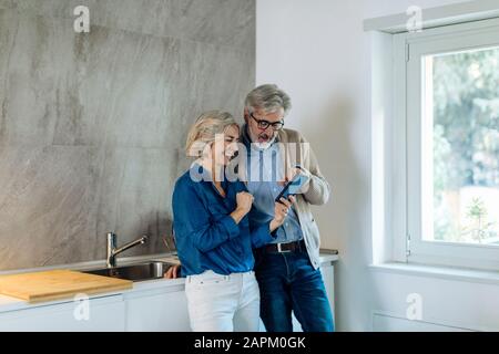 Couple d'âge mûr utilisant le téléphone cellulaire ensemble dans la cuisine à la maison Banque D'Images