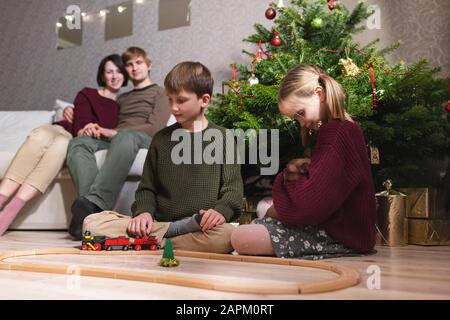 Garçon et fille avec catt, jouant avec un train jouet sur le chemin de fer en bois près de l'arbre de Noël et les parents assis sur le canapé derrière leurs enfants Banque D'Images