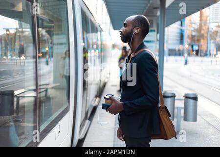 Un jeune homme d'affaires avec du café se tient à l'arrêt de tram pour écouter de la musique avec des écouteurs et un smartphone Banque D'Images