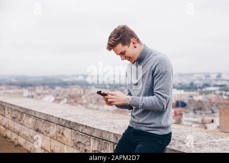 Blond jeune homme avec un pull écrivant un message sur son téléphone mobile tout en souriant à Lleida Banque D'Images