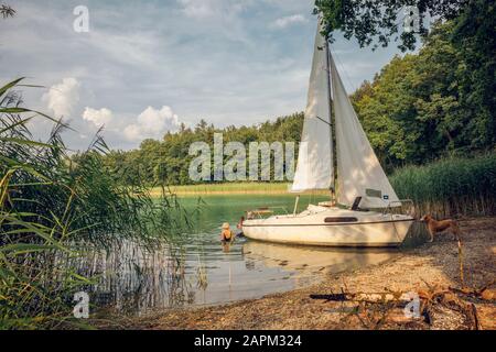 Bateau à voile au bord du lac, chien et deux personnes nageant Banque D'Images