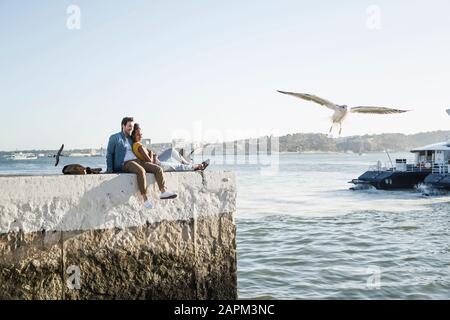 Jeune couple assis sur la jetée au bord de l'eau en profitant de la vue, Lisbonne, Portugal Banque D'Images