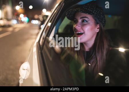 Femme assise sur l'arrière d'une voiture dans la ville la nuit, en regardant hors de la fenêtre de voiture Banque D'Images