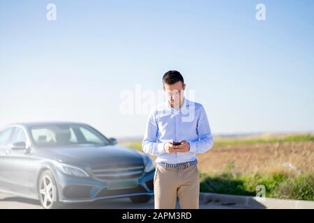 Jeune homme d'affaires avec voiture sur la route de pays en utilisant le smartphone Banque D'Images