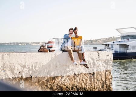 Un jeune couple heureux assis sur la jetée au bord de l'eau en prenant un selfie, Lisbonne, Portugal Banque D'Images