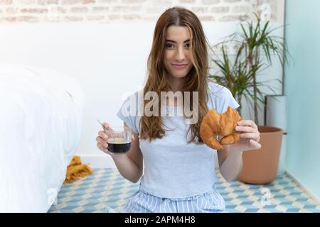 Jeune femme assise sur le sol et tenant une tasse de café et un croissant Banque D'Images